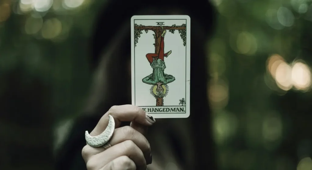 A modern witch holding 'The Hanged Man' Tarot card, which symbolizes magickal gnosis